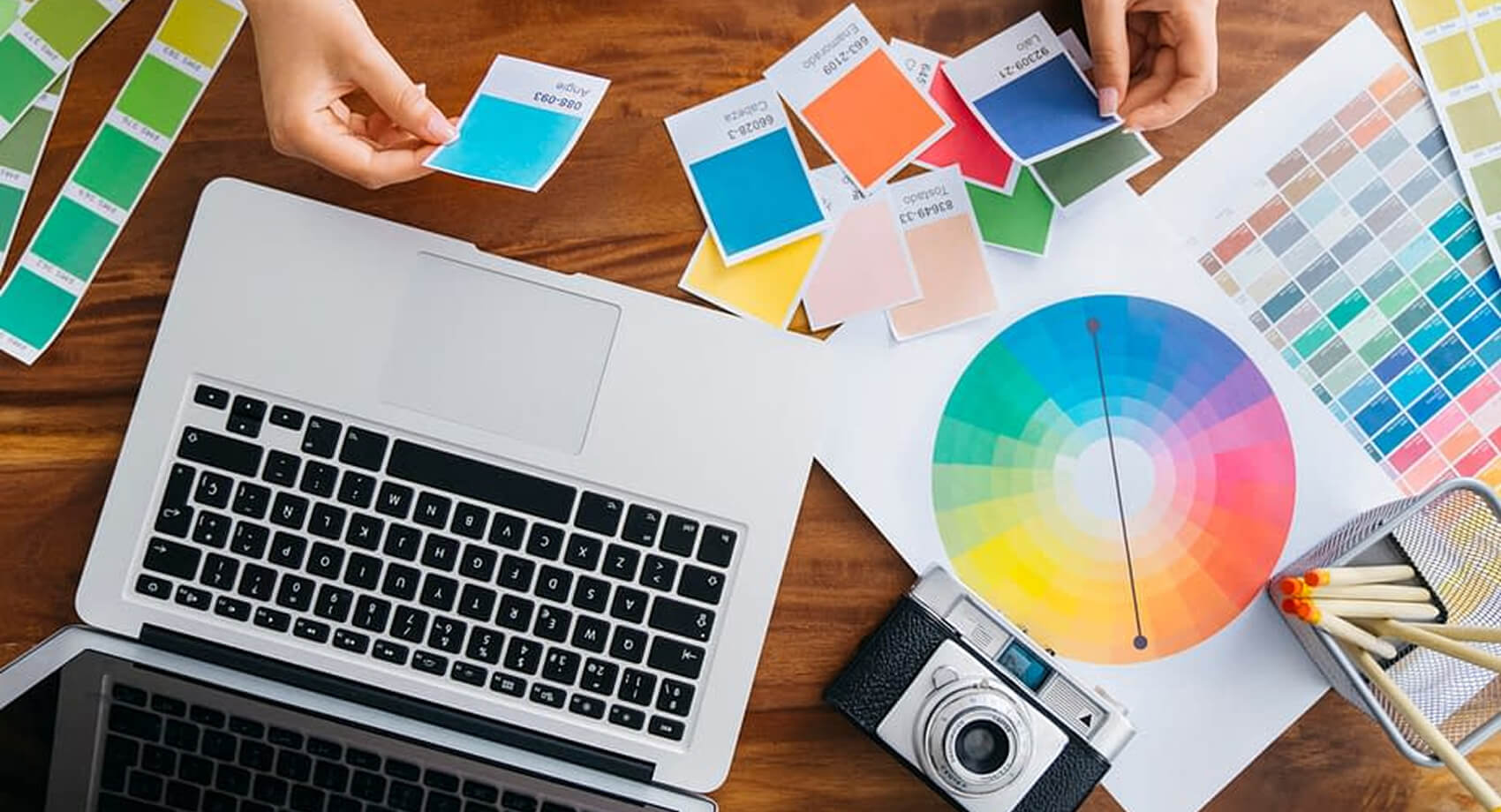 Handen met kleurstalen, een laptop, een kleurenwiel, stiften en een camera – de perfecte hulpmiddelen voor het ontwerpen van Unieke Websites.