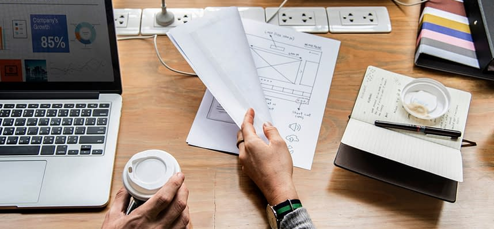 Iemand die achter een bureau zit met een laptop, een koffiekopje, een notitieboekje en documenten, en aan een project werkt.